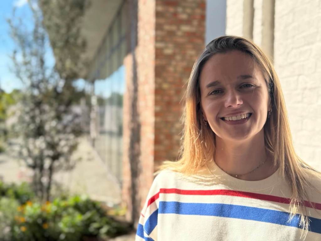 Hier zie je een lachende Evelien Nieste in een gestreept shirt, staand voor een gebouw met grote ramen en een groene omgeving.